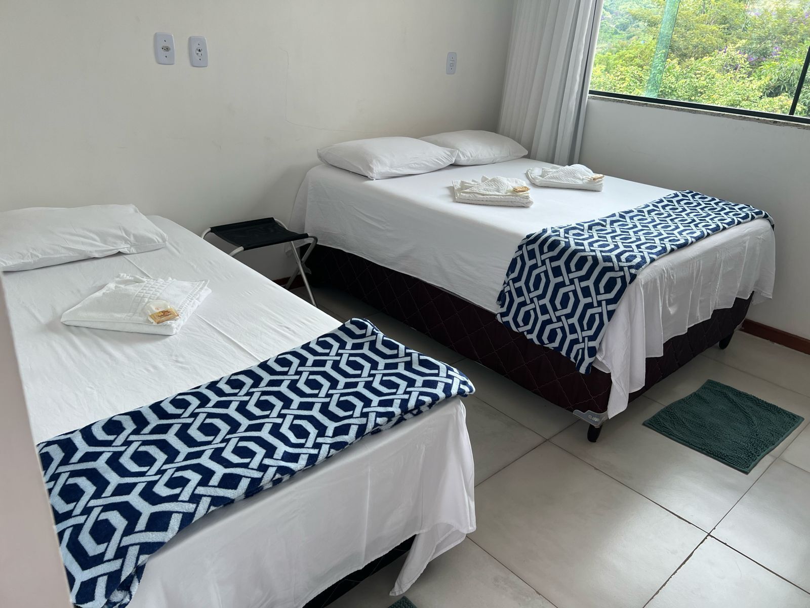 Deux lits simples avec du linge de lit blanc et des couvertures à motifs, une petite table et une fenêtre donnant sur un paysage verdoyant.