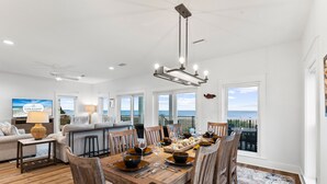 A spacious dining area with a wooden table set for six, surrounded by wooden chairs. A large window offers a view of the ocean. - Angler's Nook: Stunning Beachfront Home, Private Pool, Pet-Friendly, Sleeps 12 (Indian Pass)