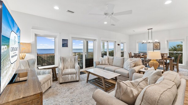 A spacious living room with a large sofa set, a coffee table, and a TV on a wooden stand. There are multiple windows offering views of the ocean. - Angler's Nook: Stunning Beachfront Home, Private Pool, Pet-Friendly, Sleeps 12 (Indian Pass)
