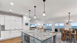A spacious kitchen with granite countertops, white cabinetry, and a large island. There is a dining area with a wooden table and chairs, and a view of the ocean through large windows. - Angler's Nook: Stunning Beachfront Home, Private Pool, Pet-Friendly, Sleeps 12 (Indian Pass)