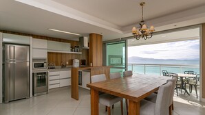 Een moderne keuken met roestvrijstalen apparatuur, een houten eettafel met stoelen en uitzicht op zee door grote ramen.