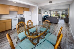 Eine moderne Küche mit Holzschränken, ein Essbereich mit Glastisch und Korbstühlen sowie ein Wohnzimmer mit Sofa und Blick ins Freie.