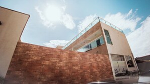 Un edificio moderno con balcón de cristal, pared de ladrillo en primer plano y cielo despejado.