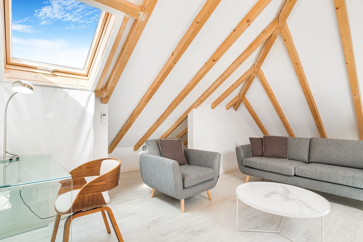 Une chambre de style loft moderne avec un plafond en pente, des poutres en bois, une grande lucarne, un canapé gris, un fauteuil gris, une table basse blanche et un bureau en verre avec une chaise.