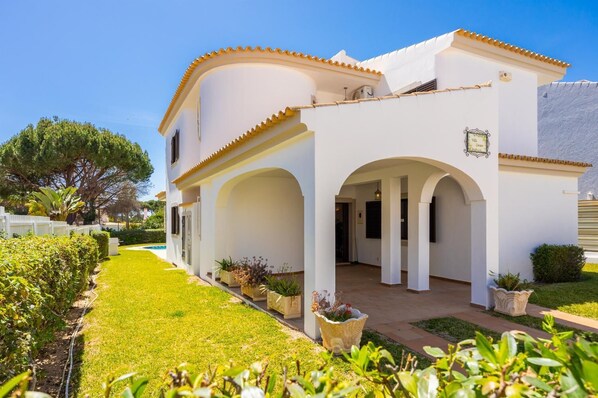 A white building with arches and a tiled roof, surrounded by a well-maintained garden and a clear sky. - Villa Cavaco - Four Bedroom Villa, Sleeps 8 (Quarteira)