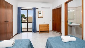 A hotel room with two beds, a wooden dresser, a bathroom with a sink and mirror, and a view through a glass door. - Villa Cavaco - Four Bedroom Villa, Sleeps 8 (Quarteira)