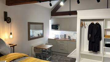 A compact kitchen with a microwave, sink, and cabinets. There is a dining table with chairs and a small seating area with a chair and a pillow. A mirror hangs on the wall, and a coat rack with two robes is visible.