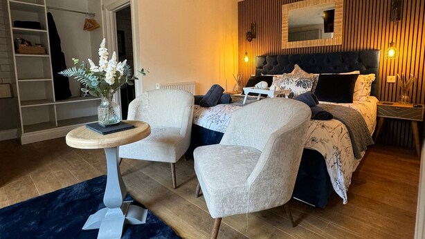 A modern bedroom with a bed, two armchairs, a small round table, and a large mirror. - The Old Stables (Northampton)