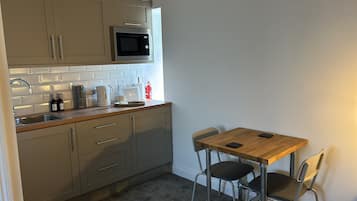 A compact kitchen with a microwave, kettle, toaster, and coffee maker. There is a small dining table with two chairs and a fire extinguisher on the wall.