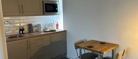 A compact kitchen with a microwave, kettle, toaster, and coffee maker. There is a small dining table with two chairs and a fire extinguisher on the wall.