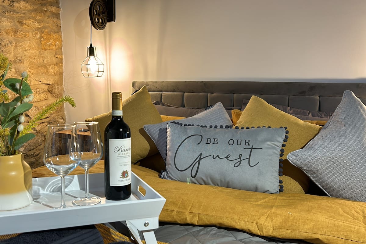 A cozy living room with a stone wall, a sofa with cushions, a tray with a bottle of wine and glasses, and a decorative pillow with the text "BE OUR Guest".