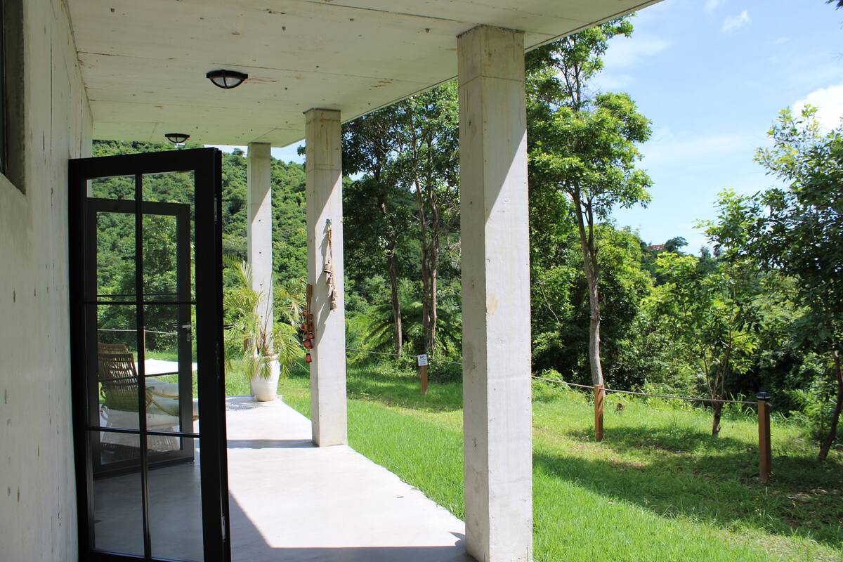 Une véranda couverte avec des piliers en béton, une porte vitrée donnant sur une pièce où se trouvent une chaise et une table, et une vue sur un paysage verdoyant et luxuriant.