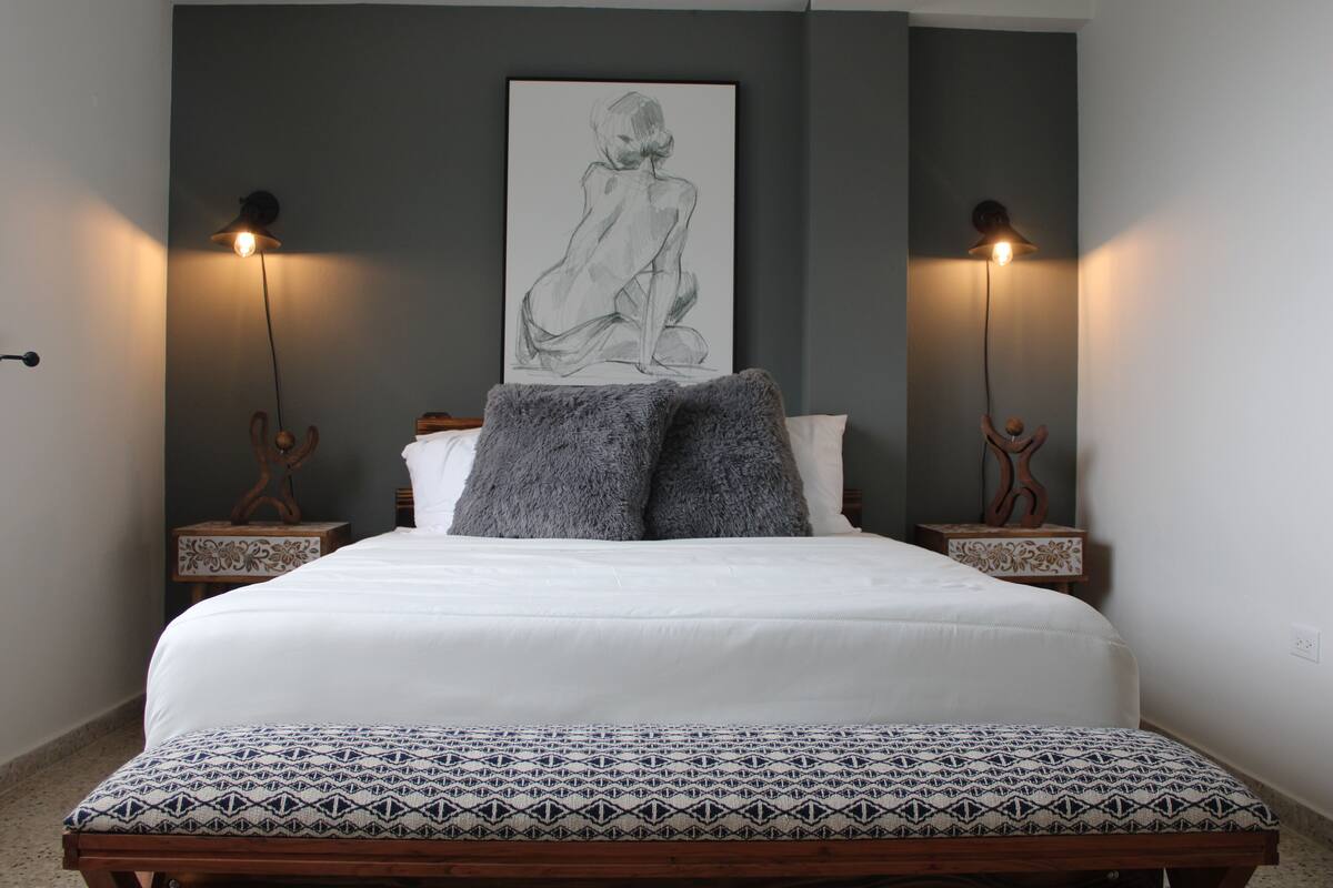 Une chambre à coucher avec un grand lit, deux tables de chevet, une lampe fixée au mur et un tableau encadré.