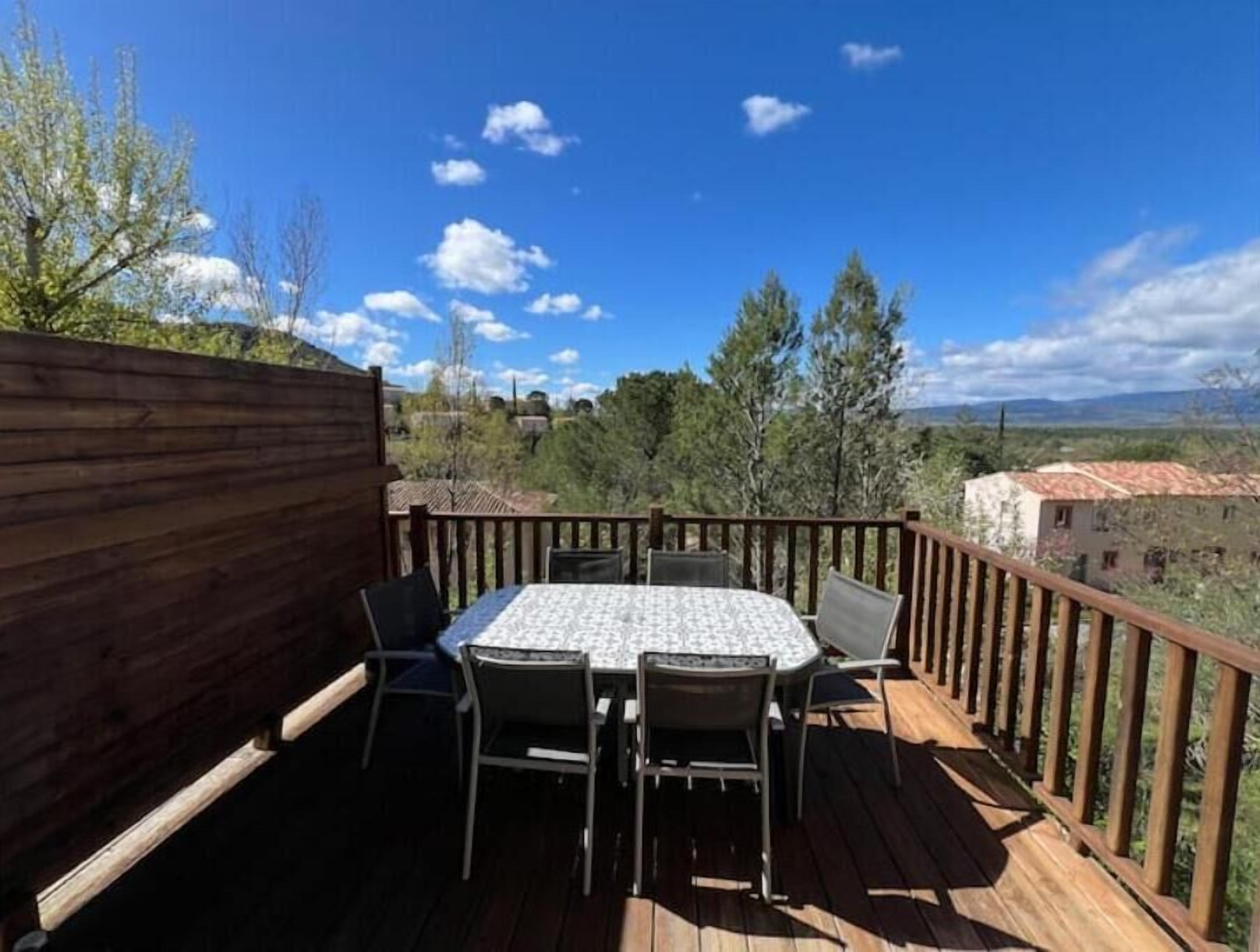 Une terrasse en bois avec une table ronde et des chaises, entourée d’une balustrade en bois, offrant une vue panoramique sur un paysage pittoresque, avec des arbres et des bâtiments.