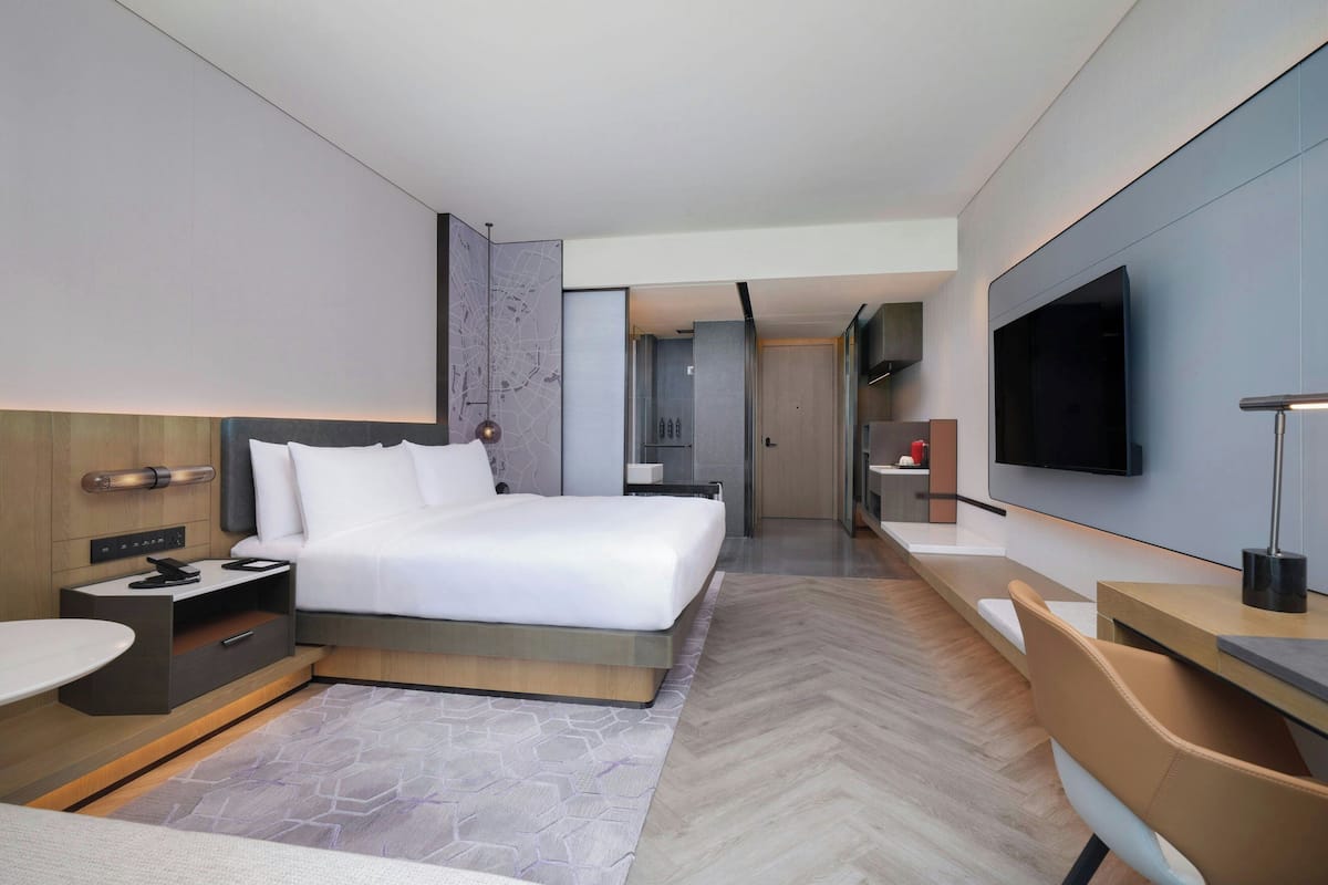A modern hotel room with a large bed, a desk, a TV, and a bathroom visible through an open door.