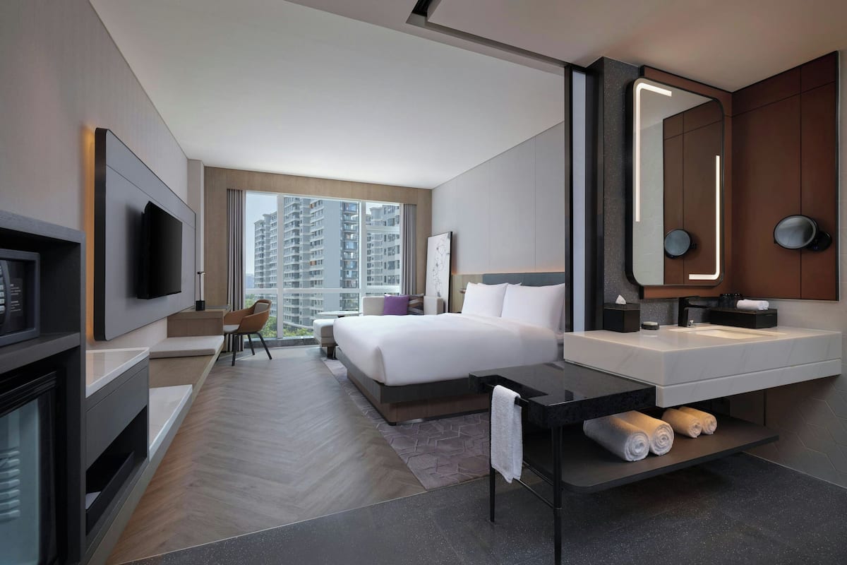A modern hotel room with a large bed, a TV, a desk, and a bathroom with a large mirror and sink.