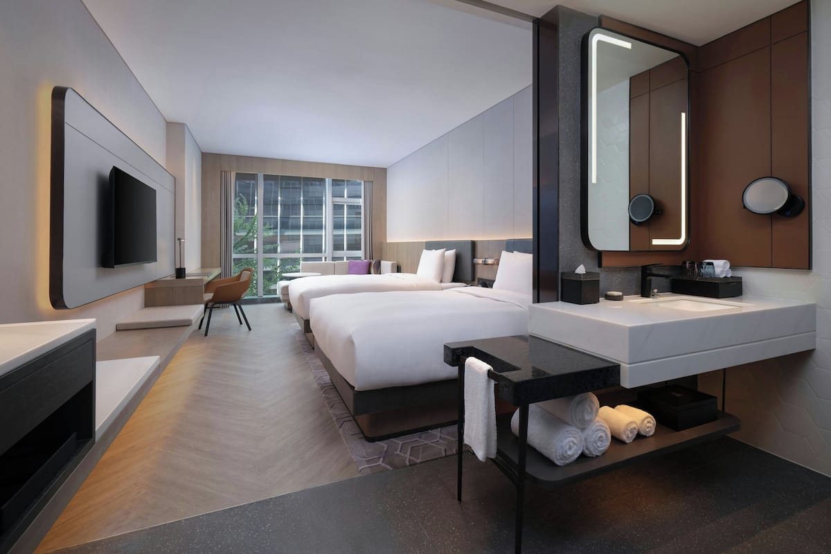 A modern hotel room with a large bed, a separate sofa, a flat-screen TV, a large mirror, a bathroom with a sink and towel rack.