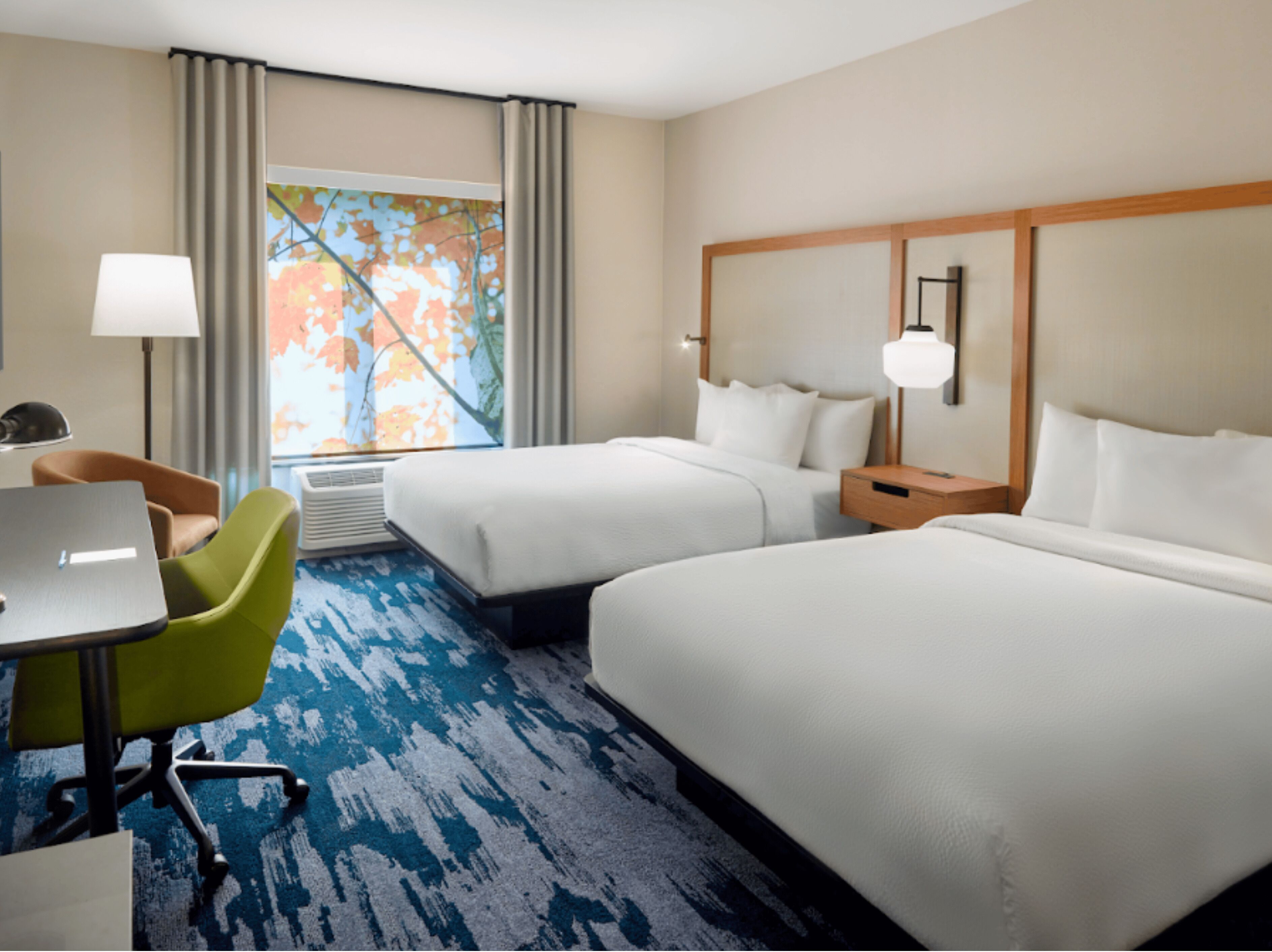 Une chambre d’hôtel avec deux lits, un bureau avec une chaise, une lampe et une fenêtre donnant sur un paysage d’automne aux couleurs flamboyantes.