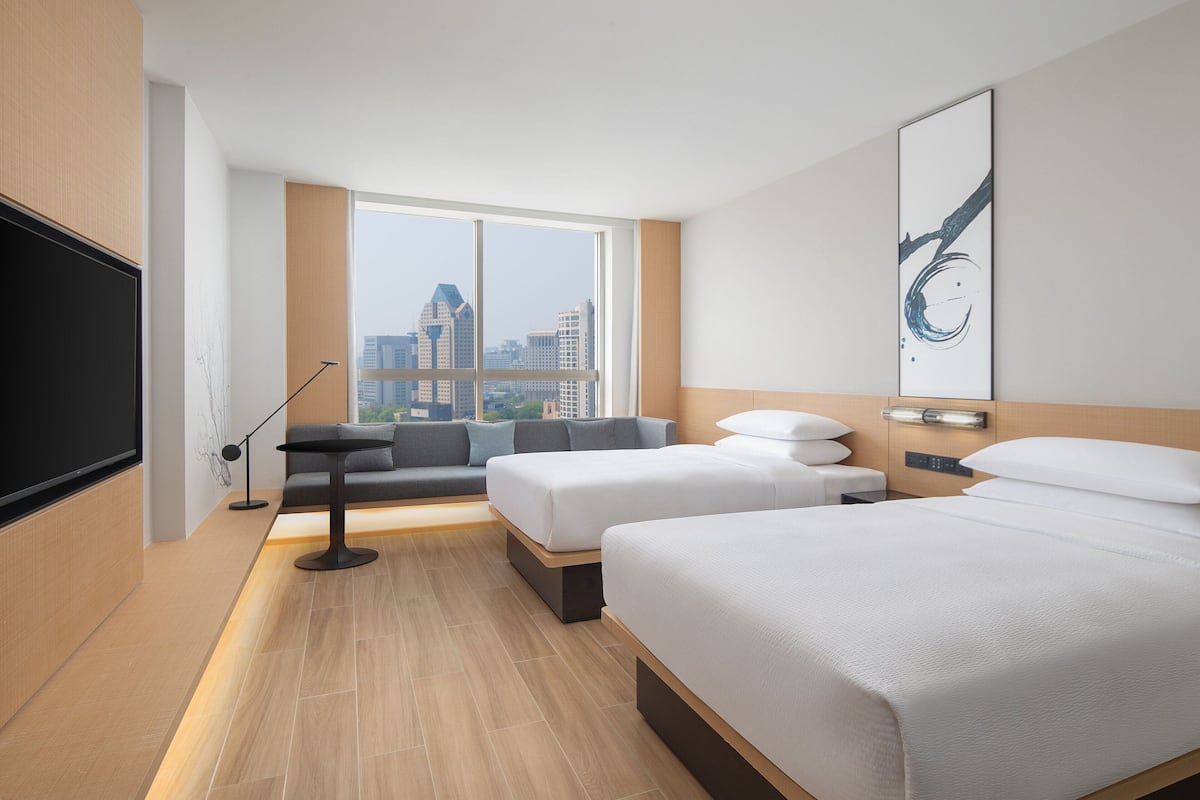 Una habitación de hotel moderna con dos camas, un área de descanso, un ventanal con vistas a la ciudad y una obra de arte colgada en la pared.