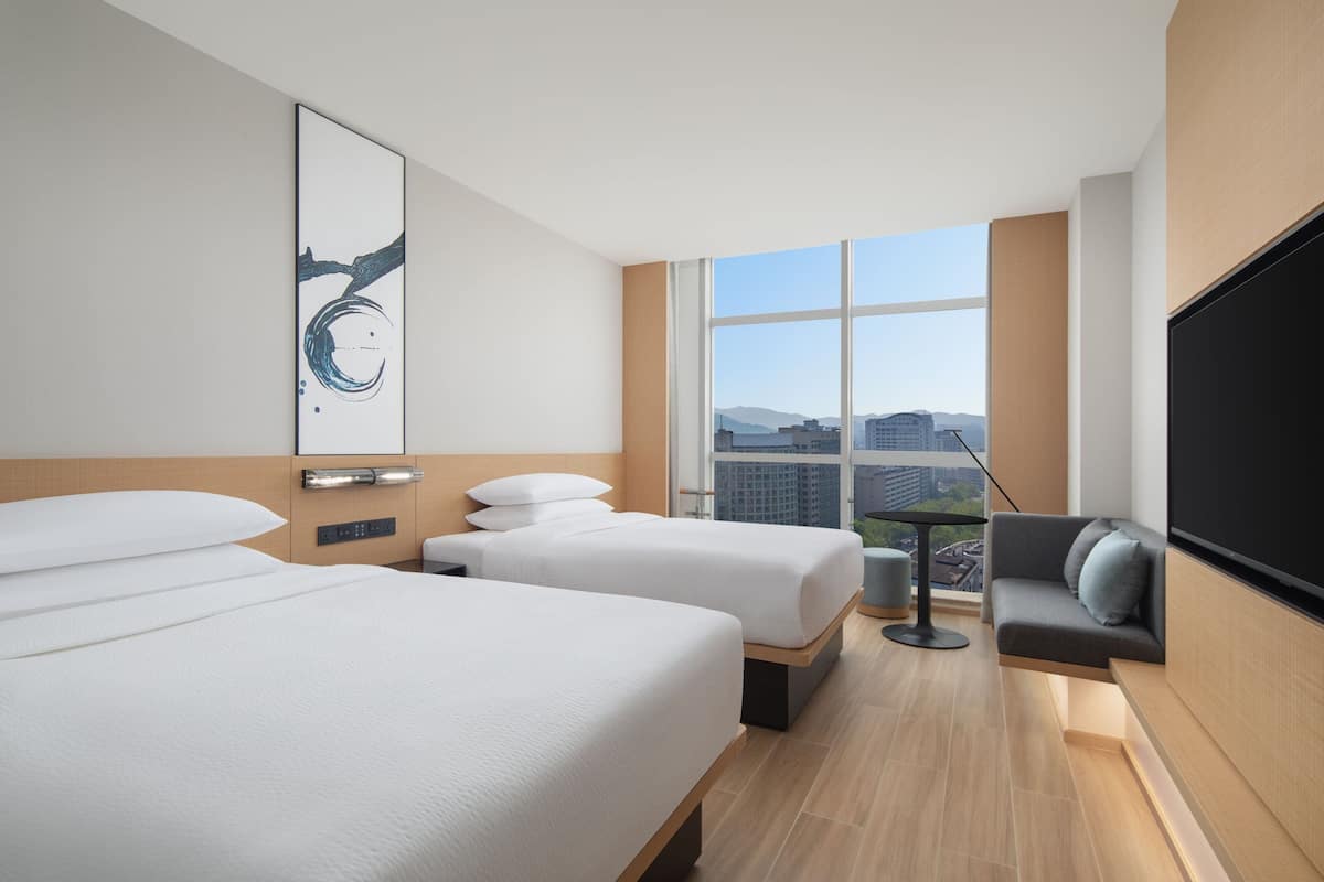 Habitación de hotel con dos camas, un sofá, un televisor y un ventanal con vistas a la ciudad.