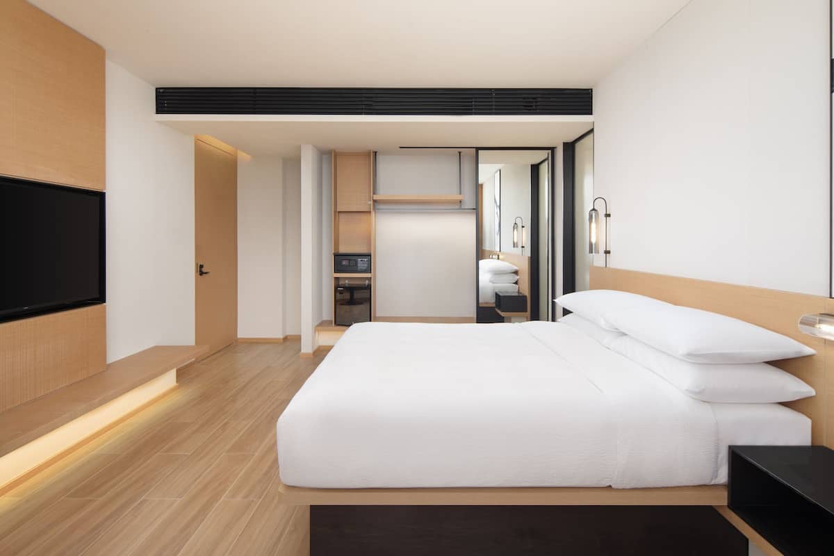 Habitación de hotel moderna con una cama grande, un televisor de pantalla plana, suelo de madera y un baño visible a través de una puerta abierta.