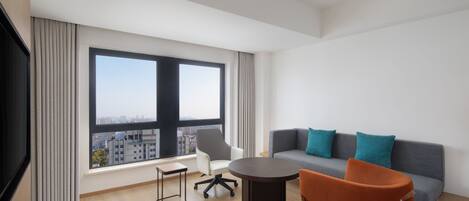 A modern living room with a large window, a grey sofa with teal pillows, a round table, a white armchair, and a wooden floor.