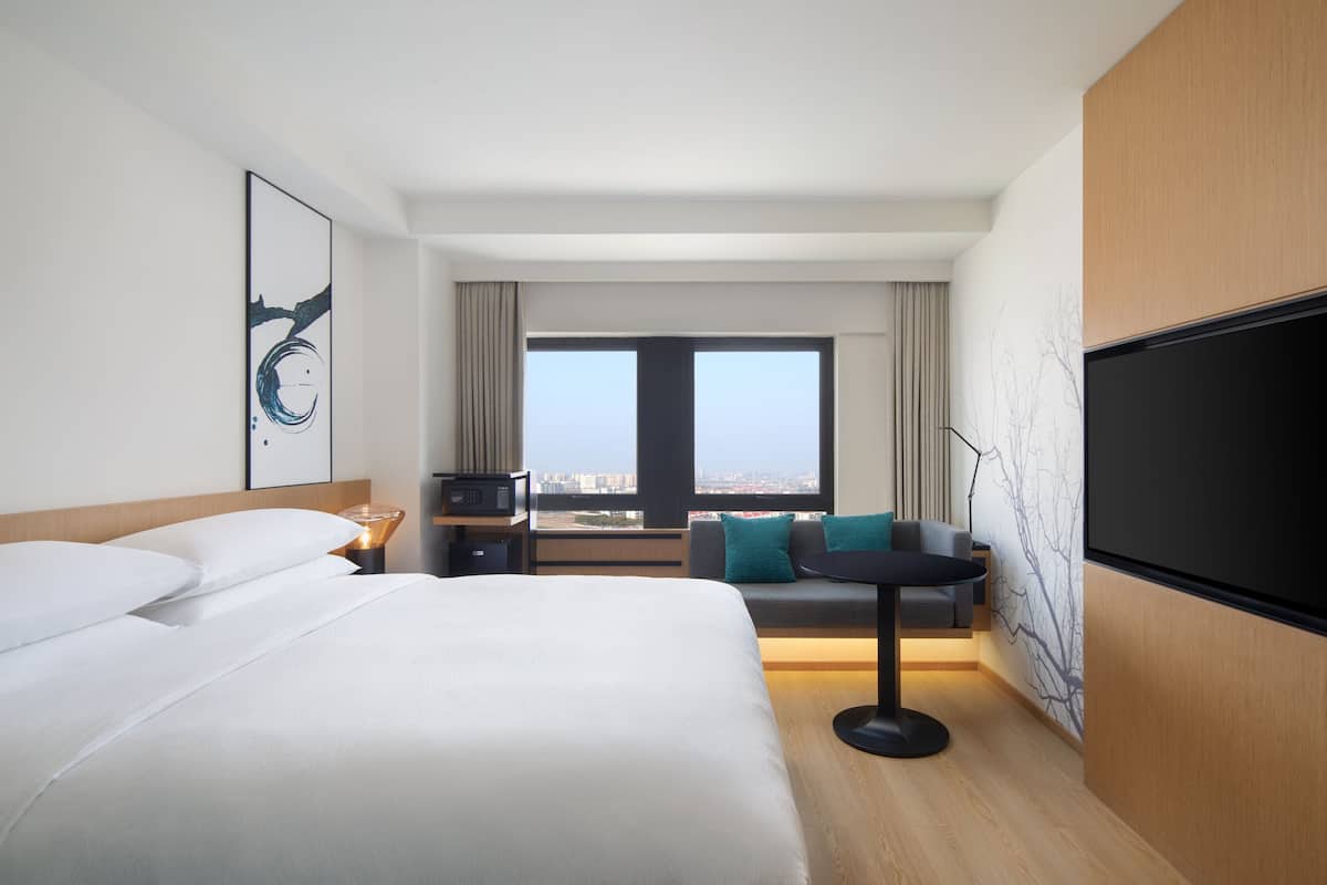 A modern hotel room with a large bed, a desk, a chair, a TV, and a view of the city.