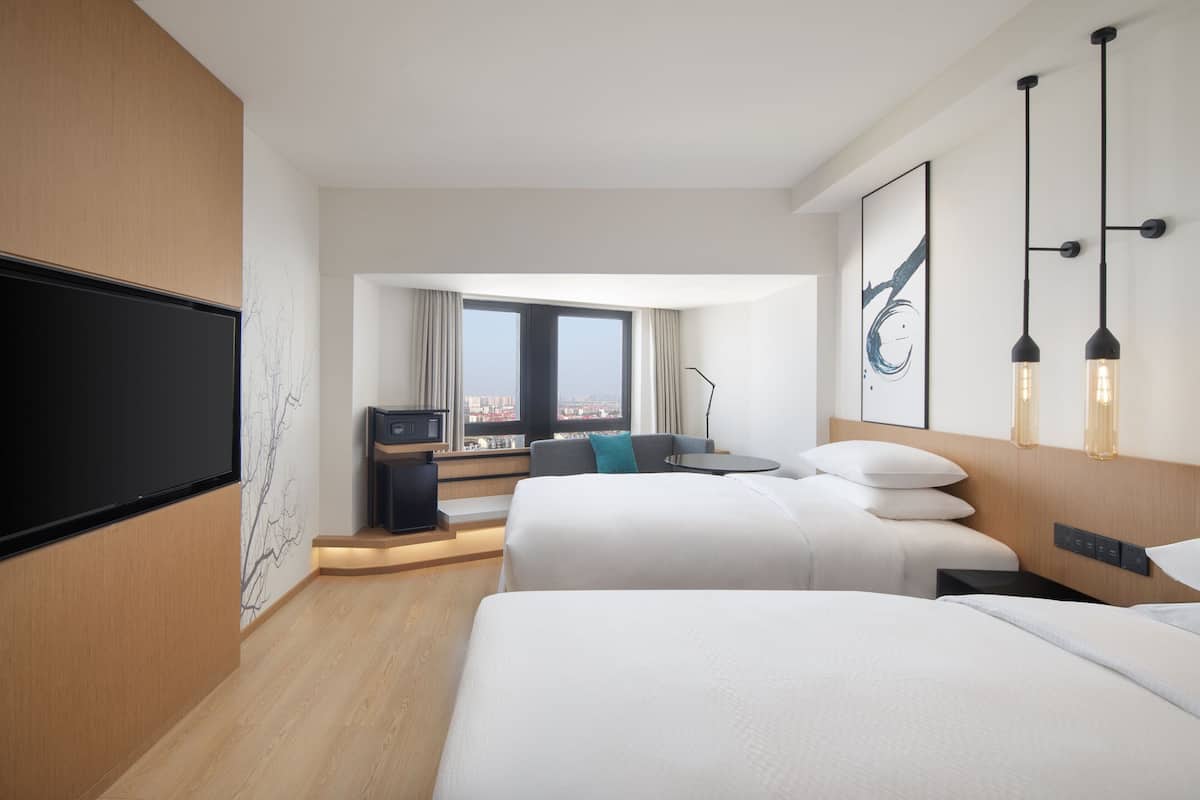 A hotel room with two beds, a desk, a TV, and a large window with a city view.