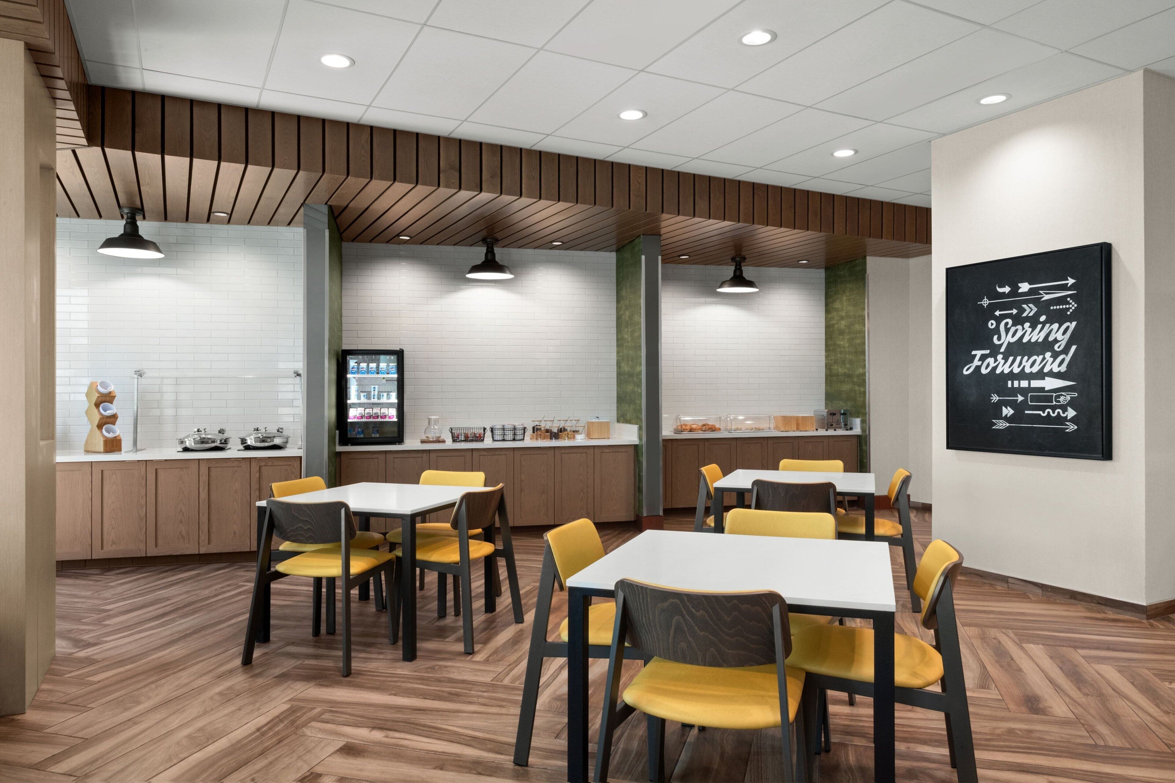 Un espace repas moderne avec un plancher en bois, des chaises jaunes et des tables blanches. On y trouve un comptoir de cuisine avec divers objets, ainsi qu’un grand panneau mural sur lequel on peut lire : « Avancez l’heure ».