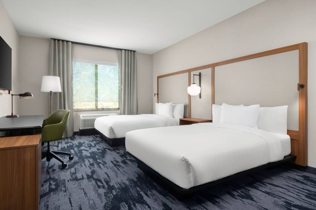 Habitación de hotel con dos camas, un escritorio con silla, una lámpara y un ventanal grande con cortinas.