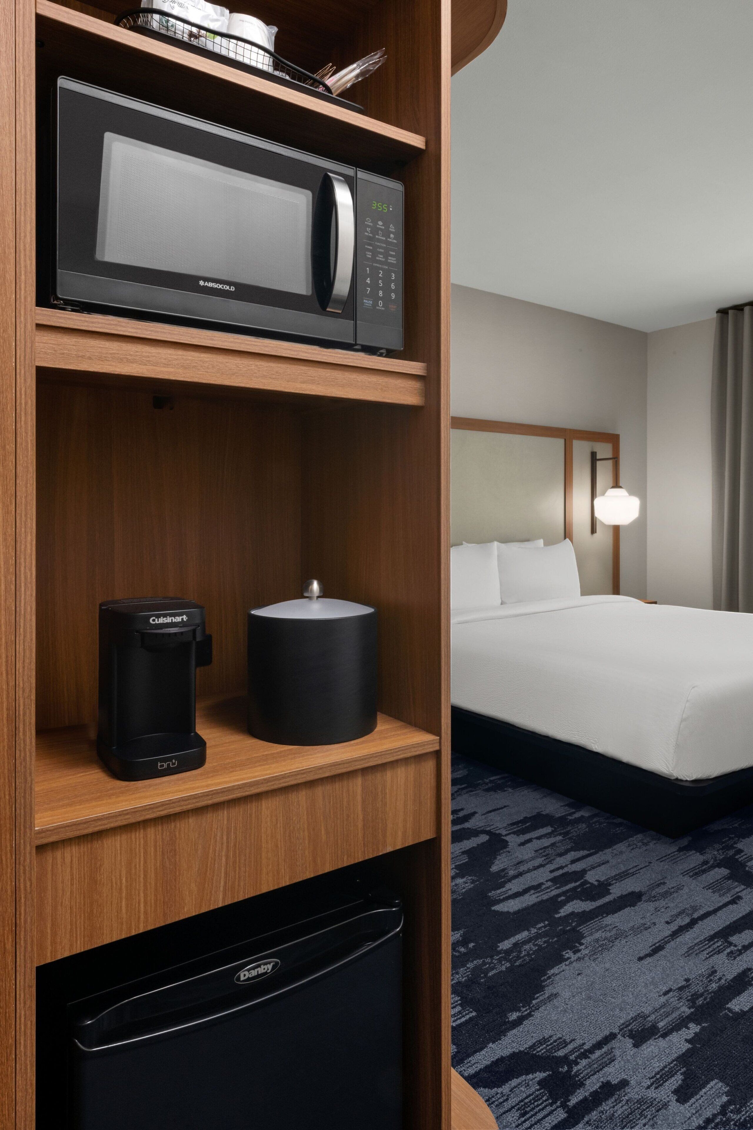 Habitación de hotel con microondas, cafetera y lavavajillas en un estante, una cama con ropa blanca y una lámpara de noche.