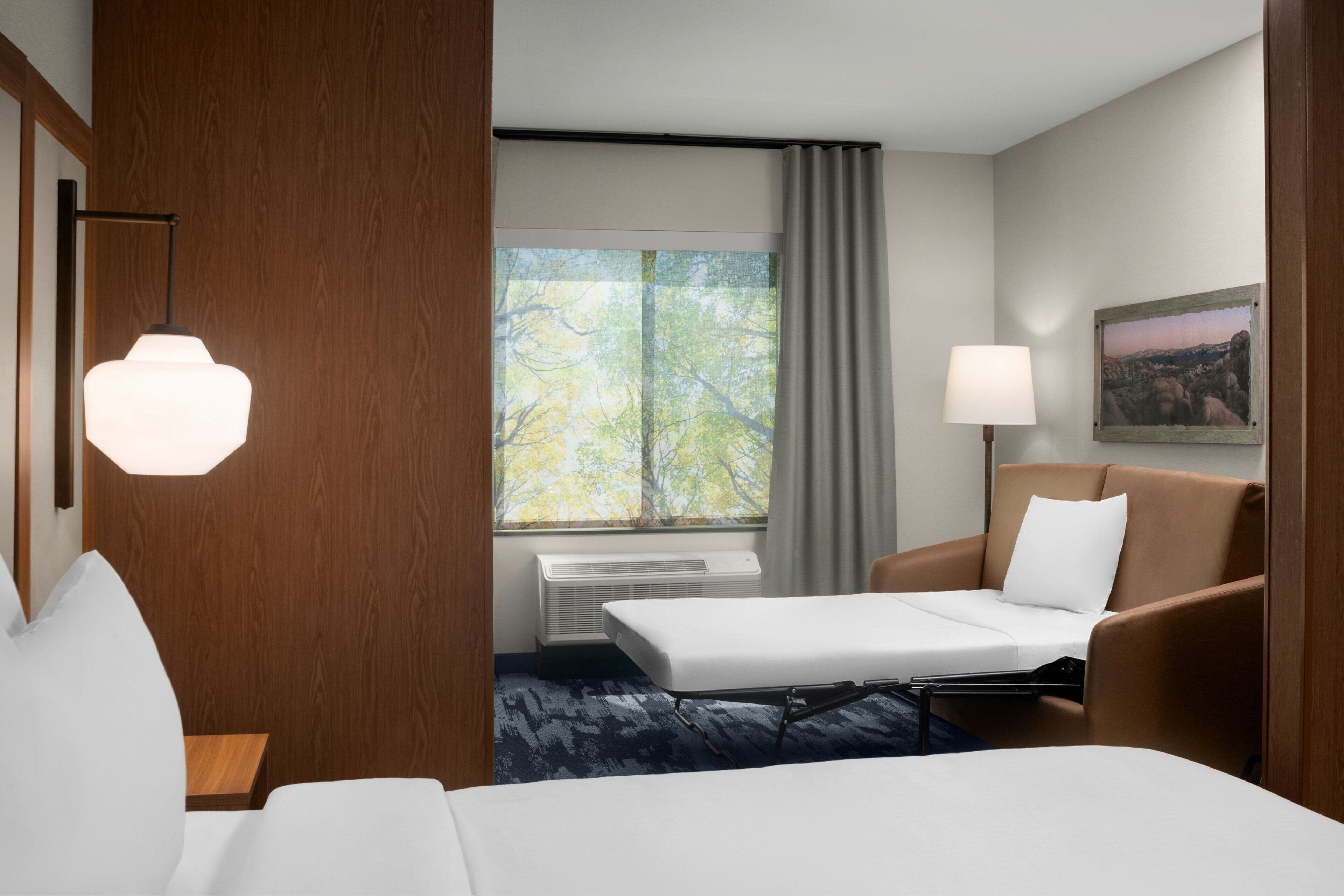 A hotel room with a bed, a sofa, a desk, a lamp, a painting, and a window with curtains.
