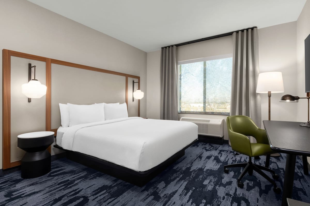 A hotel room with a large bed, a desk, a chair, a lamp, a window with curtains, and a wall-mounted mirror.