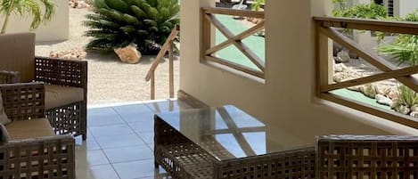 Un patio con mobiliario de mimbre y una mesa de vidrio, con vistas a un jardín con palmeras y al océano.