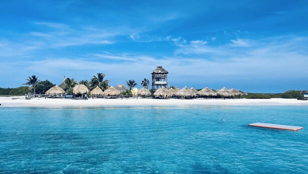 Un resort en la playa con cabañas de techo de paja, una torre central con el letrero 'Mermaid' y aguas azules cristalinas.