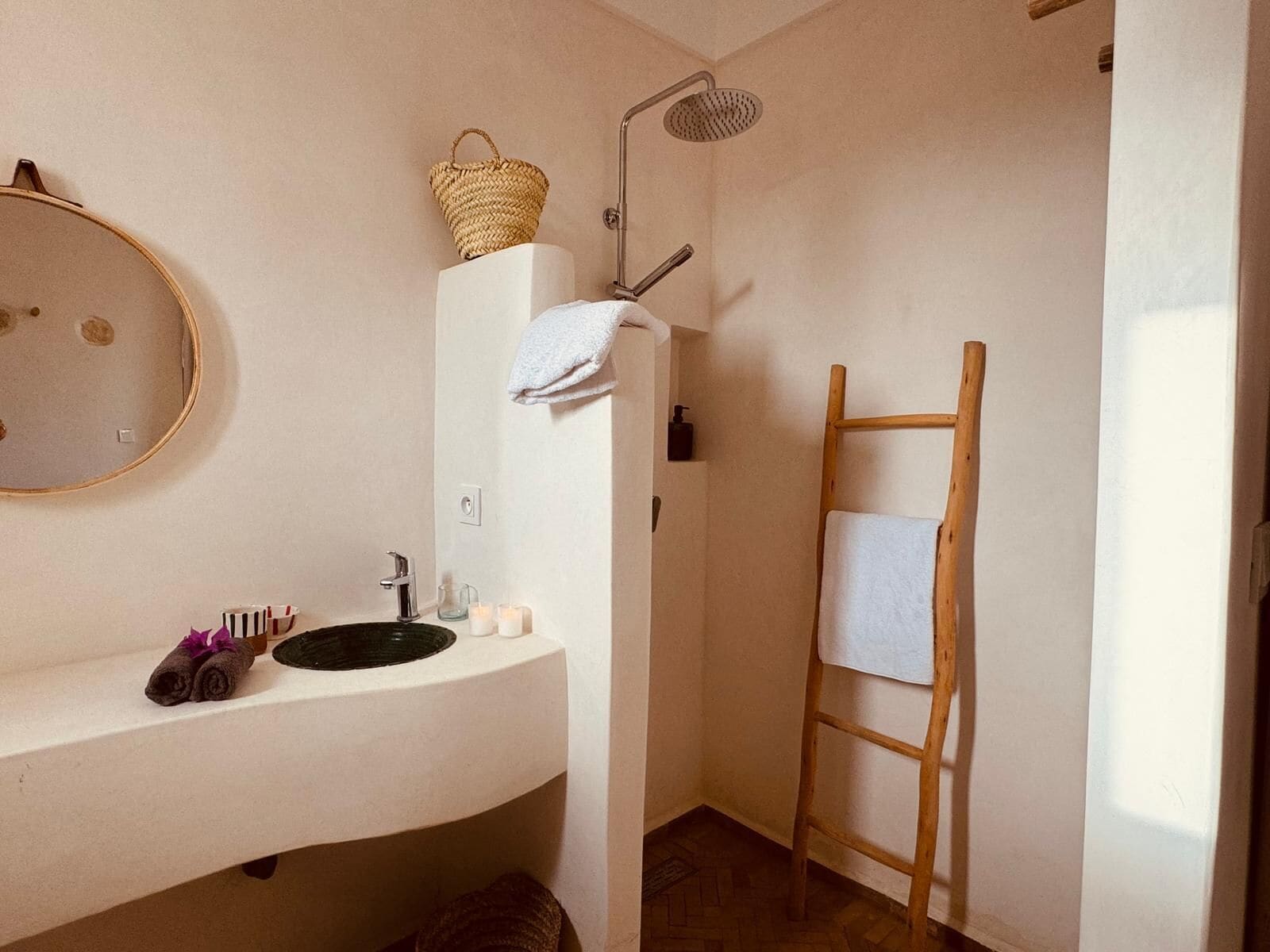 A bathroom with a round mirror, a sink with a black basin, a wooden ladder, and a shower with a handheld showerhead.