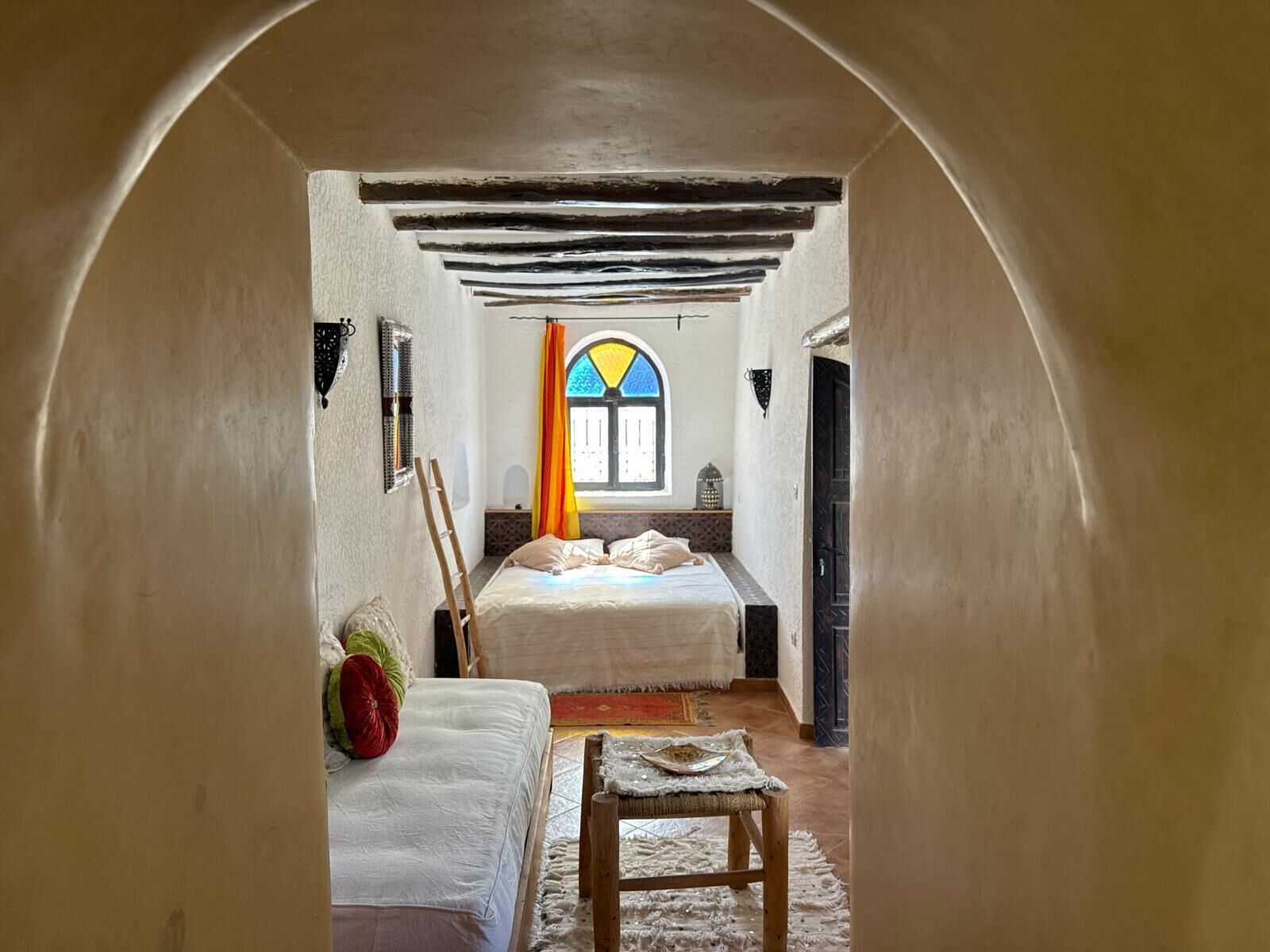 Ein schmaler Flur führt zu einem Schlafzimmer mit einem Bett, einem kleinen Tisch und einem Fenster mit einem bunten Vorhang.