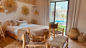 A bedroom with a bed, wooden chairs, a woven bench, and a view of a pool outside. - RIAD EUCALYPTUS, ETHNIC HOTEL (Essaouira)