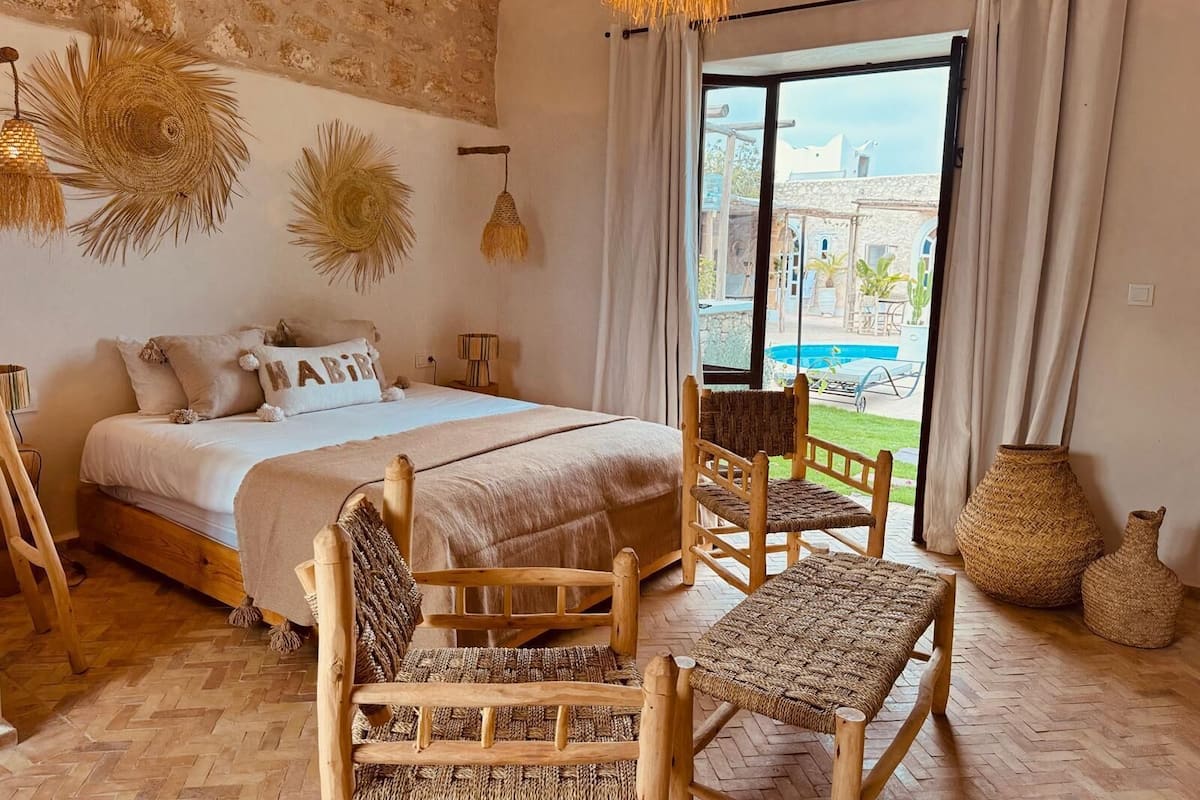 A bedroom with a bed, wooden chairs, a woven bench, and a view of a pool outside.