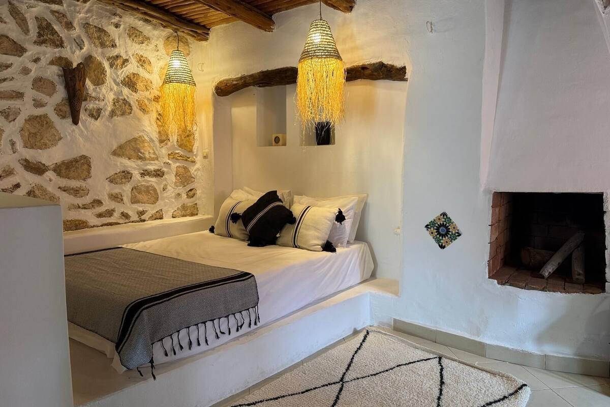 A cozy bedroom with a stone wall, a wooden ceiling, a bed with a patterned throw, and two hanging lamps.