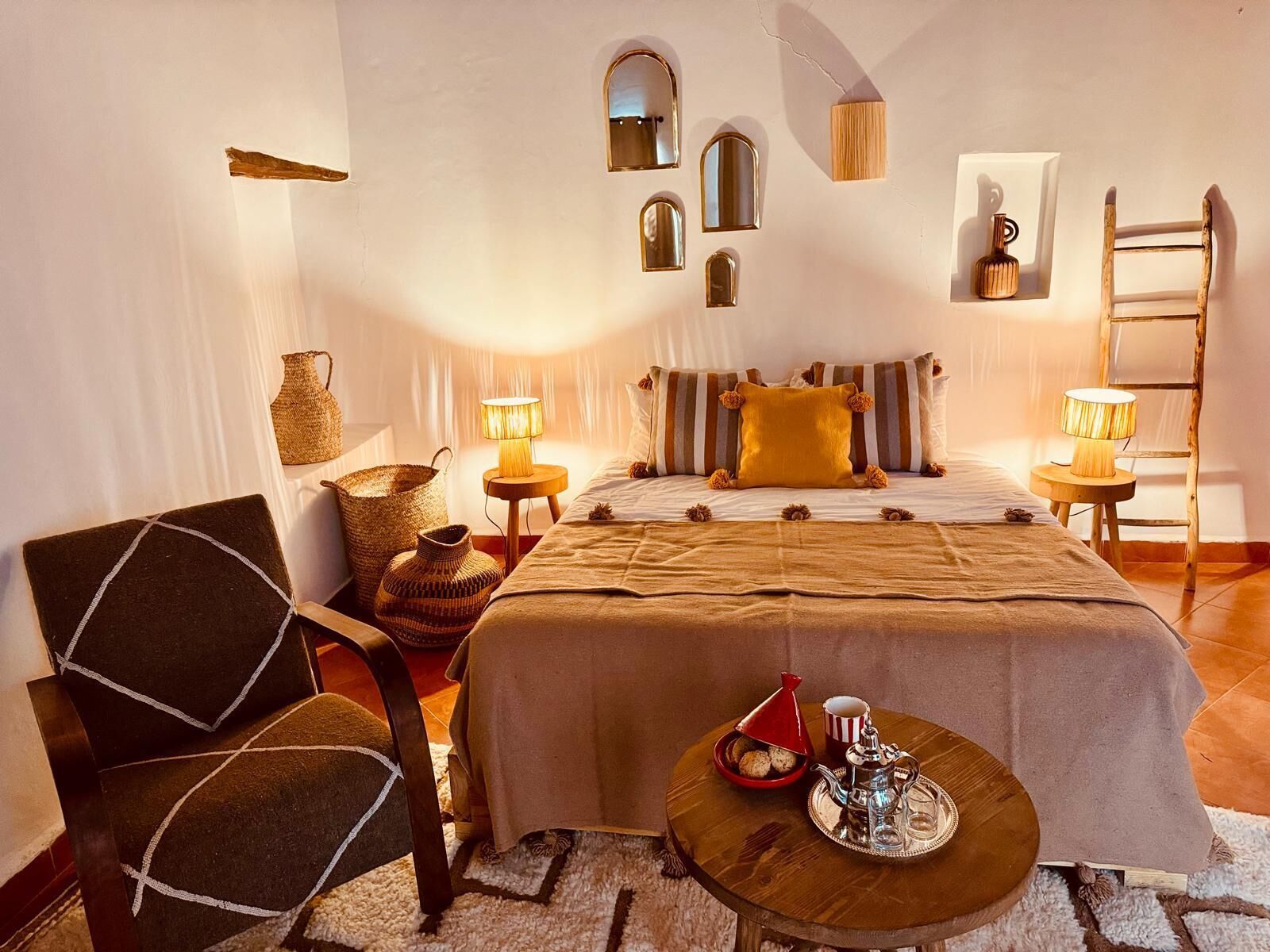 A cozy bedroom with a bed, two bedside lamps, a wooden chair, a small round table with a teapot and cookies, and a woven basket.