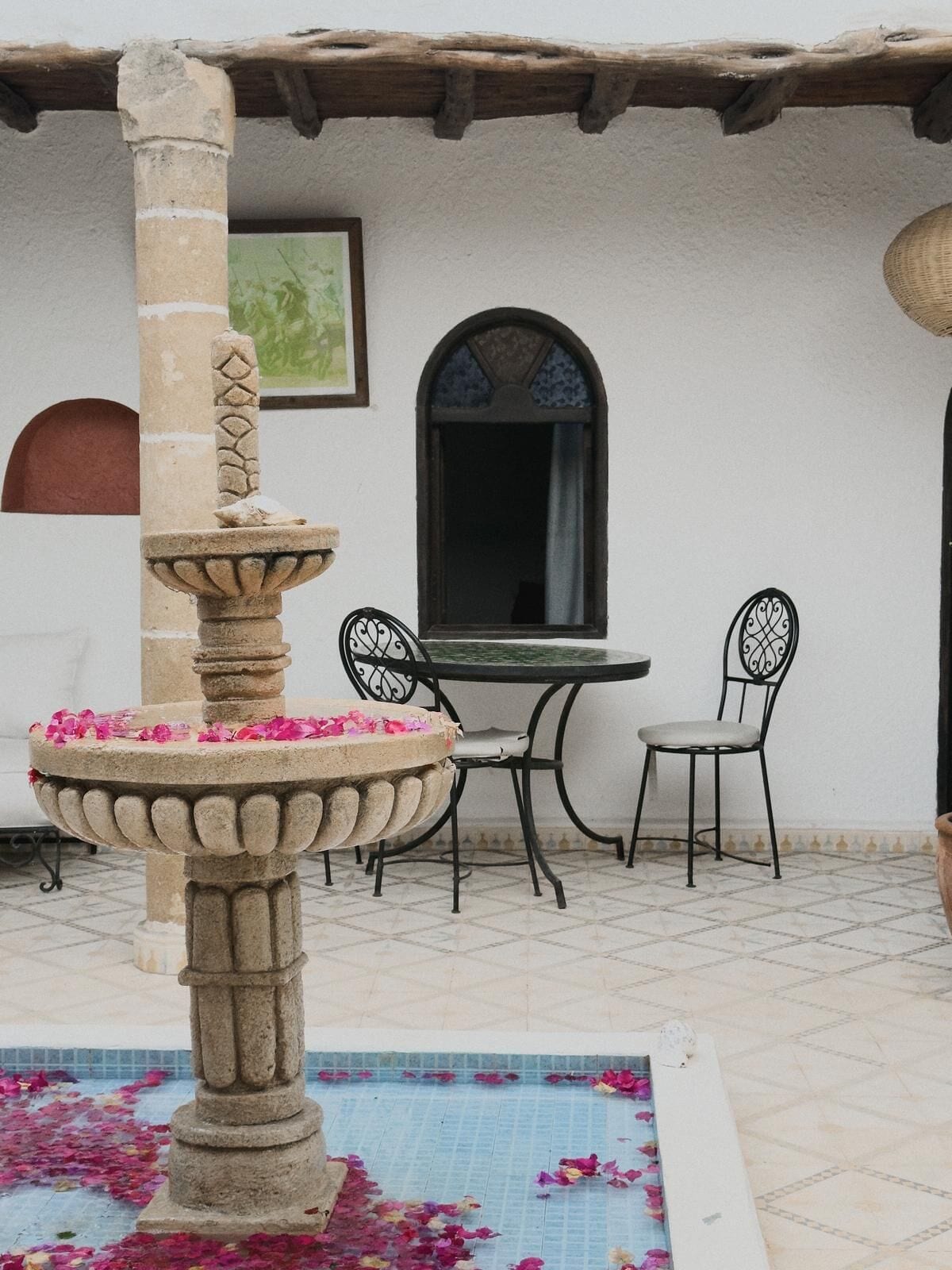 Une fontaine en pierre ornée de pétales de fleurs, une table avec des chaises et une fenêtre dotée d’un cadre décoratif.