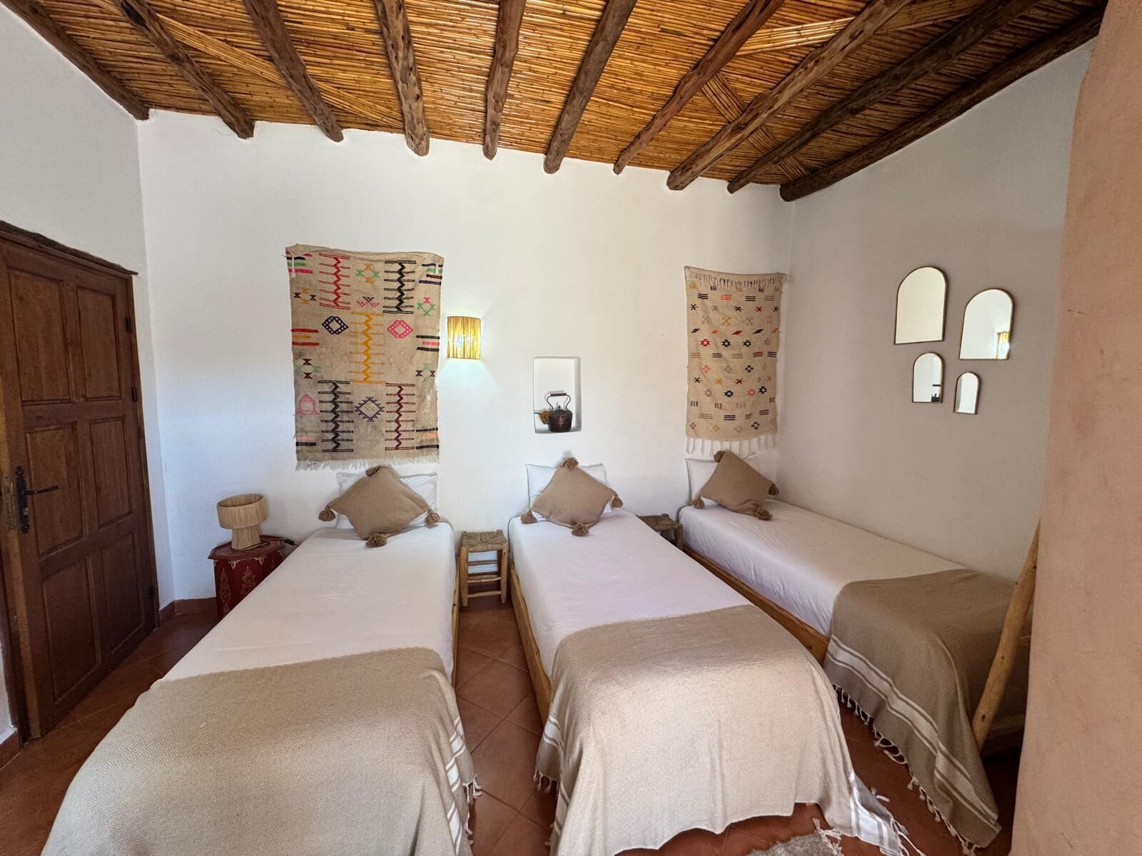A room with three beds, a wooden door, a wall with a patterned tapestry, and a small shelf with a lamp.