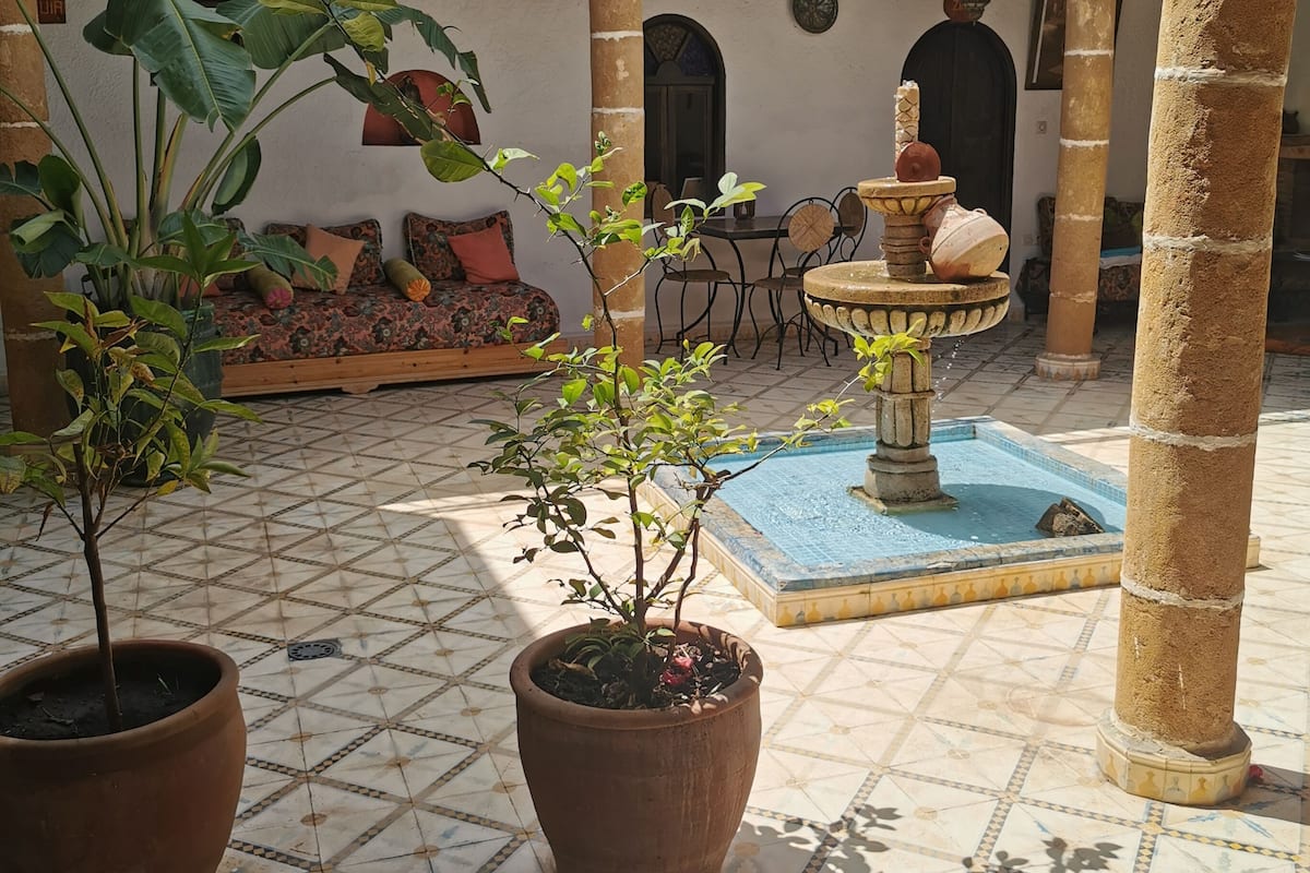 A courtyard with tiled flooring, potted plants, a fountain, and a seating area with cushions.
