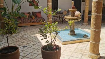 Un patio con suelo de baldosas, plantas en macetas, una fuente y una zona de estar con cojines.