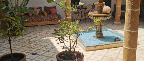 A courtyard with tiled flooring, potted plants, a fountain, and a seating area with cushions.
