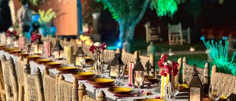 Una larga mesa de comedor al aire libre con sillas de mimbre, velas y centros de mesa florales.