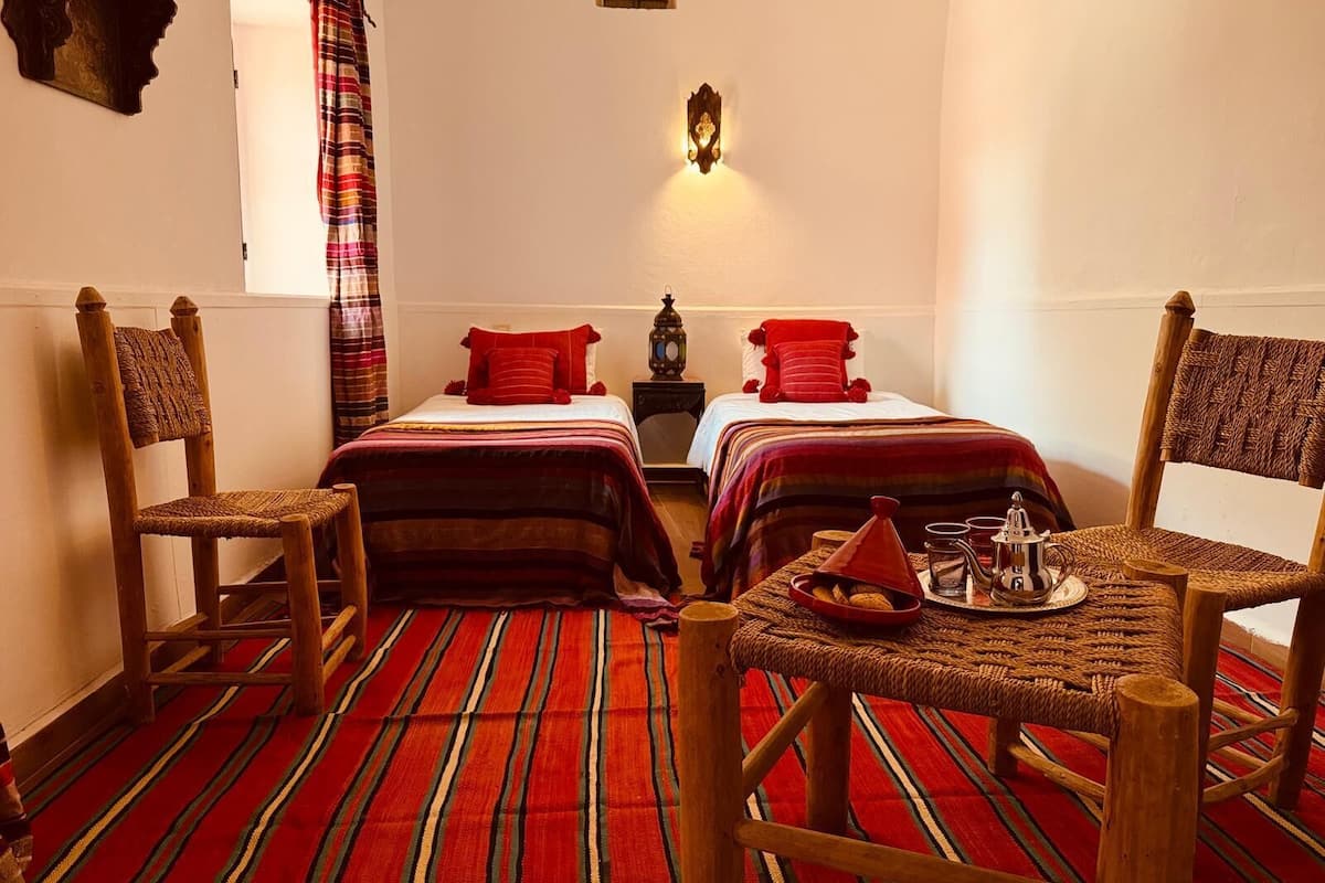 A room with two beds, a woven chair, a small table with a tray, and a wall-mounted light fixture.