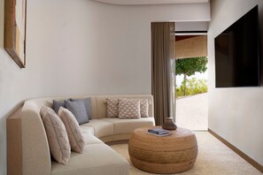 A modern living room with a beige sofa, patterned cushions, a round woven coffee table, and a flat-screen TV mounted on the wall.