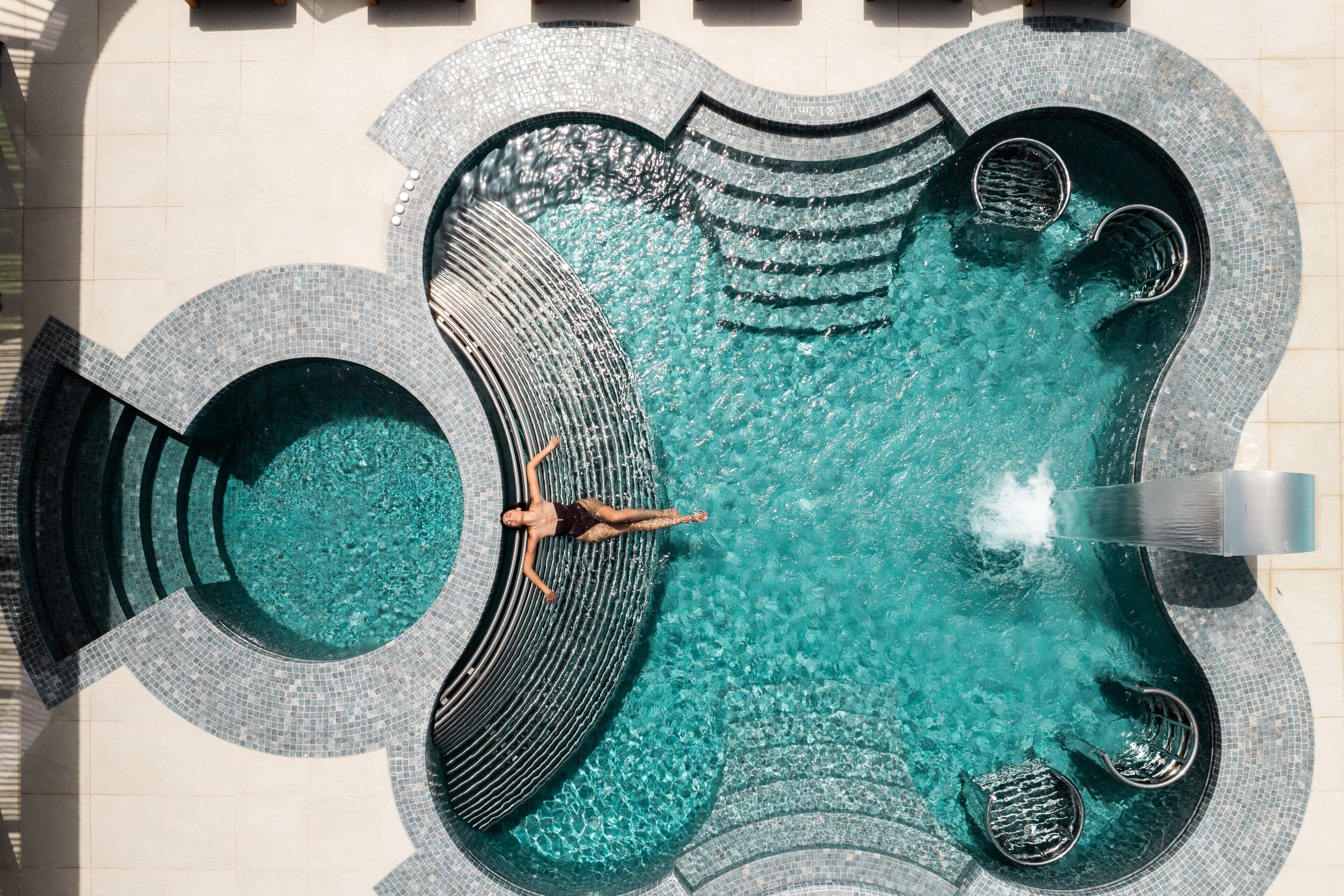 Un'ampia piscina dalla forma particolare, con diverse zone relax e una persona che si rilassa su una panca a forma di arco.